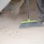 Close-up of a person's feet and the bottom of a broom, sweeping debris at a construction site, symbolizing post-construction cleaning.