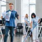 A young man with red hair holds a basket of cleaning supplies in an office, with two women cleaning in the background.